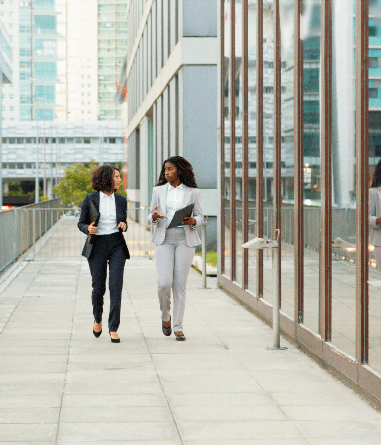 photo of two women in their 20s walking down a city sidewalk in front of a large set of windows.