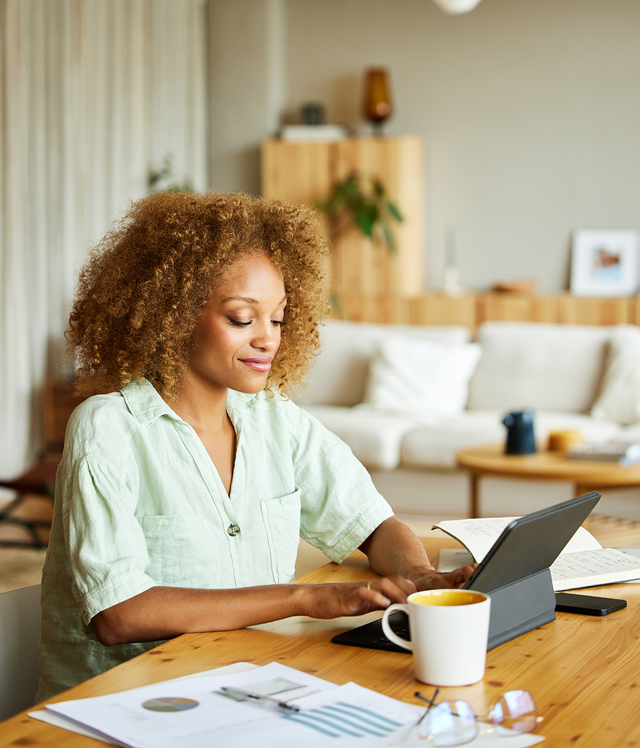Businesswoman is typing on digital tablet while sitting at desk. Female professional is working at home. She has blond curly hair.